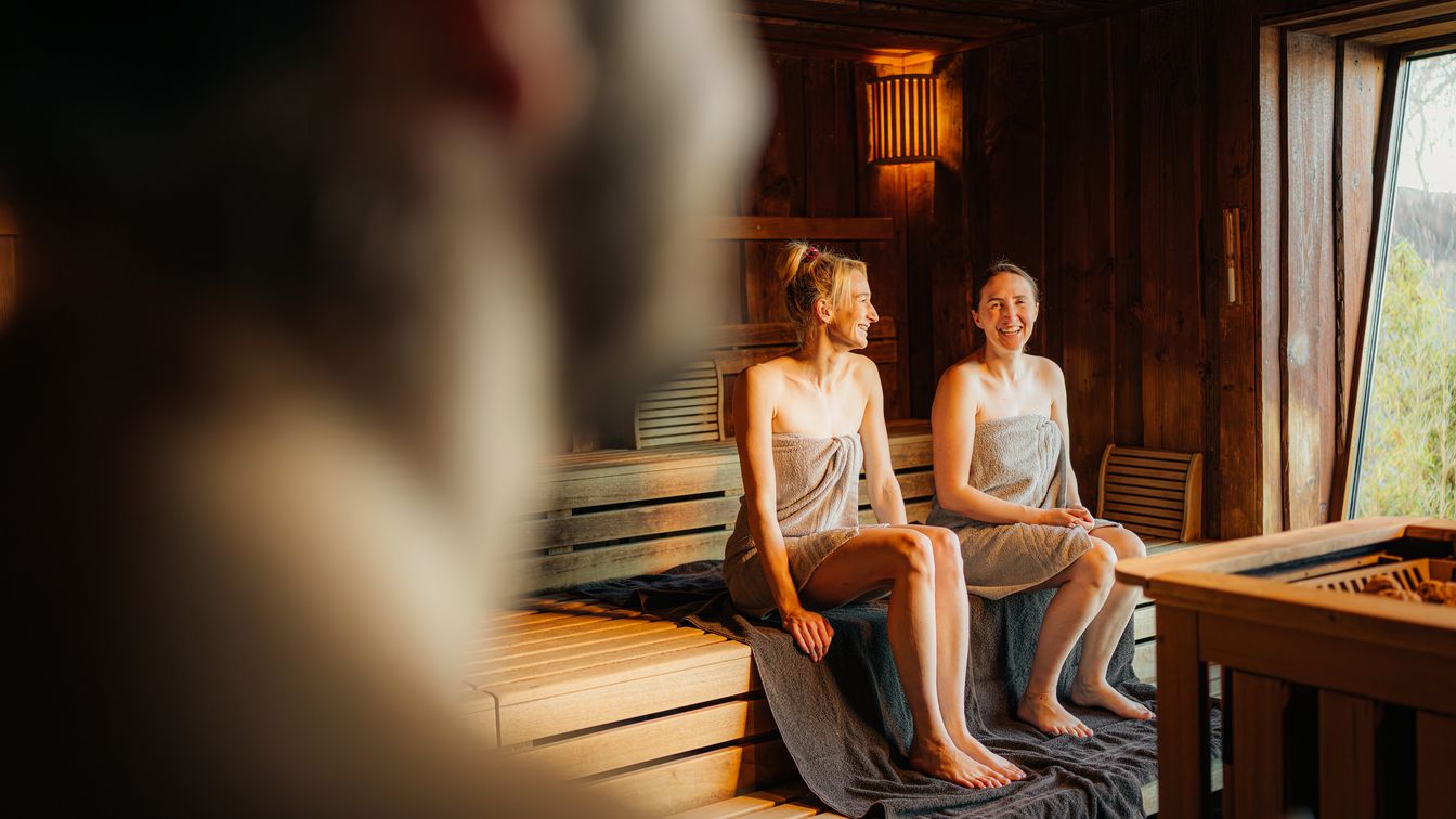Zwei Personen in Handtüchern sitzen lachend in einer holzverkleideten Sauna in der Sauna im See mit Blick nach draußen auf den Cospudener See.