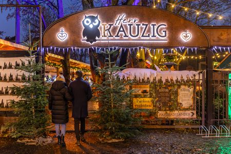 Zwei Personen betreten den Eingang zum Wintermarkt bei Herrn Kauzig, links und rechts vom Eingang stehen zwei Tannenbäume
