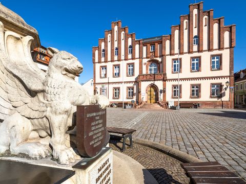 Das Bild zeigt den Marktplatz in Mügeln. Links sieht man den Brunnen und im Hintergrund das rot-weiße Rathaus der Kleinstadt.