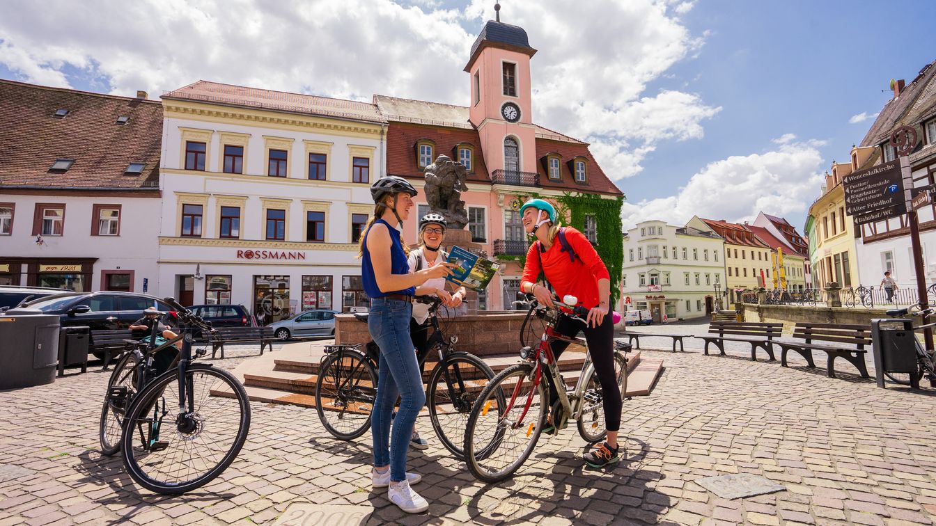 Das Bild zeigt drei Radfahrer:innen auf dem Marktplatz Wurzen, im Hintergrund sieht man die Fassade des Rathauses der Kleinstadt.