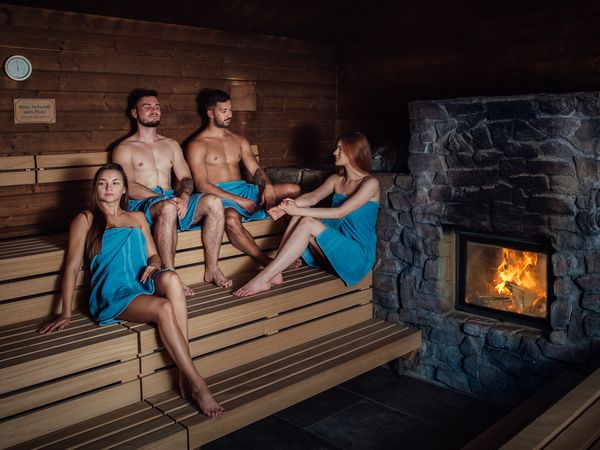 Saunieren in der Meri Sauna am Kulkwitzer See, zu sehen sind zwei Frauen und zwei Männer mit blauen Handtüchern in einer Sauna, im Kamin lodert das Feuer © Meri Sauna