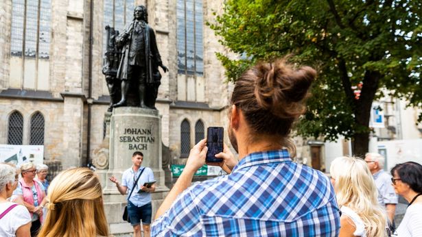 Ein junger Mann und eine junge Frau stehen während einer Stadtführung am Bach-Denkmal an der Thomaskirche Leipzig und hören einem Gästeführer zu, der Mann macht ein Foto mit seinem Smartphone, Gruppenführung, Musikstadt Leipzig, Komponisten, Johann Sebastian Bach