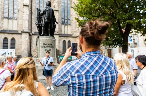 Ein junger Mann und eine junge Frau stehen während einer Stadtführung am Bach-Denkmal an der Thomaskirche Leipzig und hören einem Gästeführer zu, der Mann macht ein Foto mit seinem Smartphone, Gruppenführung, Musikstadt Leipzig, Komponisten, Johann Sebastian Bach