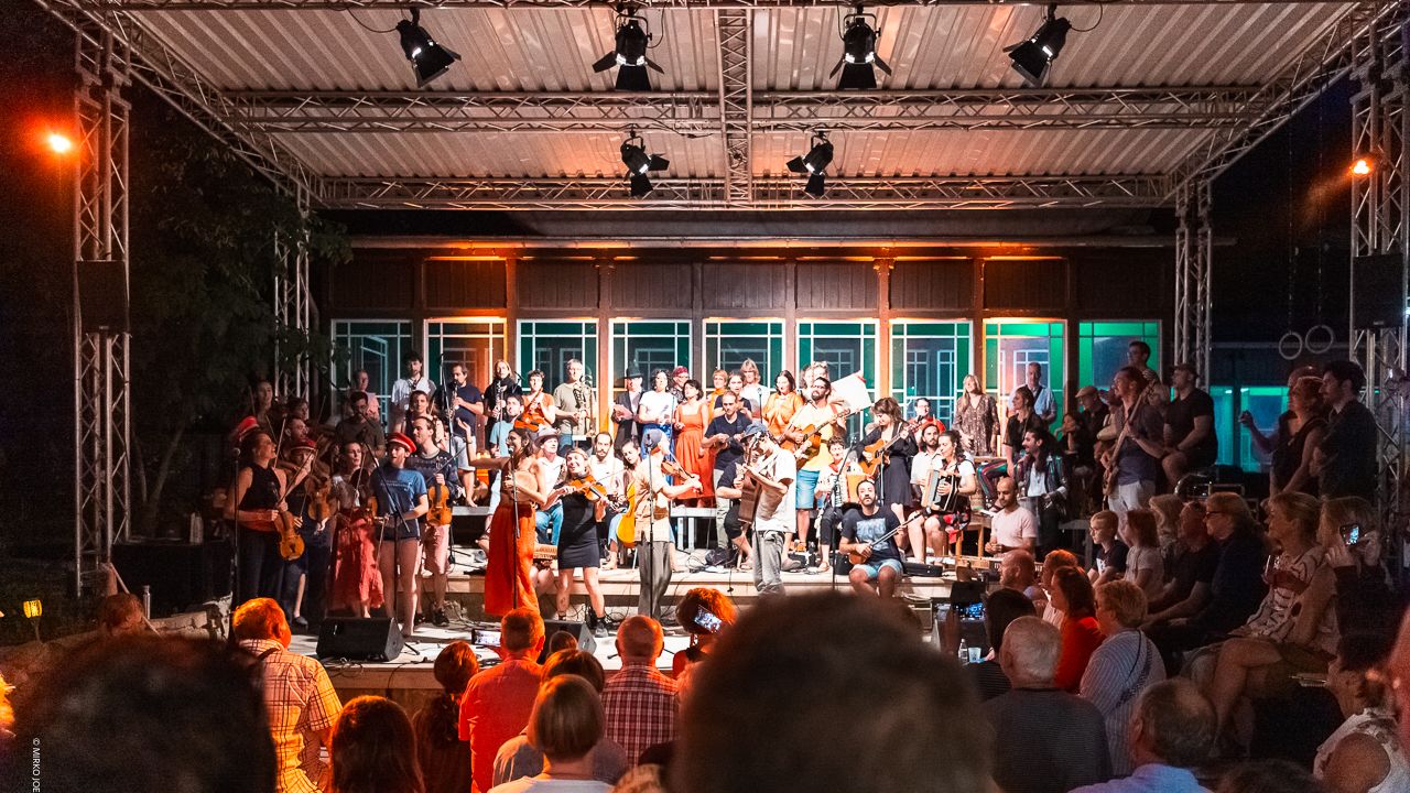 Zahlreiche Musiker mit unterschiedlichen Instrumenten stehen auf der Bühne vor dem Bahnhofsgebäude, die Atmosphäre ist locker und fröhlich