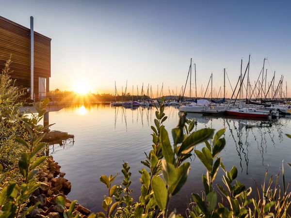Ein Teil des Störmtaler See mit einem kleinen Yachthafen im Hintergrund. Links ist eine moderne Unterkunft und im Hintergrund geht die Sonne unter. Grüne Pflanzen am Ufer rahmen das Bild.