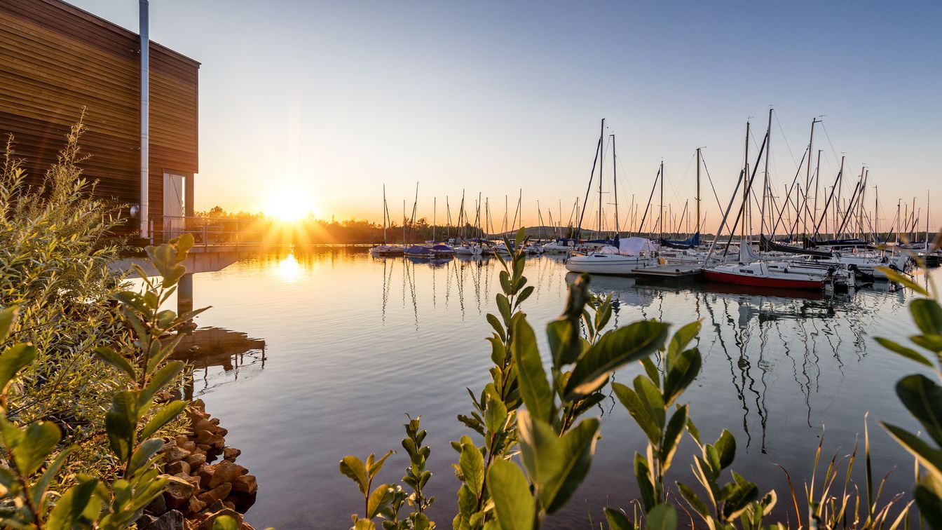 Ein Teil des Störmtaler See mit einem kleinen Yachthafen im Hintergrund. Links ist eine moderne Unterkunft und im Hintergrund geht die Sonne unter. Grüne Pflanzen am Ufer rahmen das Bild.