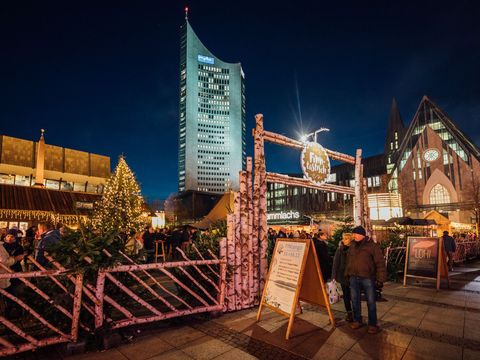 Wejście na fiński Rynek Bożonarodzeniowy i przynależne stragany na placu Augustusplatz w Lipsku, wieczorem; w tle wieżowiec City i Kościół Uniwersytecki