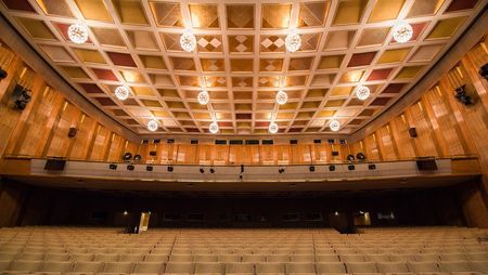 Der Saal der Oper Leipzig strahlt in goldbraunen Tönen, an der moderne Kastendecke hängen kristallene Kugellampen.