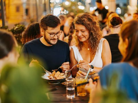 Ein junger Mann und eine junge Frau sitzen beim Abendessen am Tisch. Sie sitzen im Außenbereich eines Restaurants. Es ist schon dunkel, aber es ist atmosphärisch und gemütlich beleuchtet.
