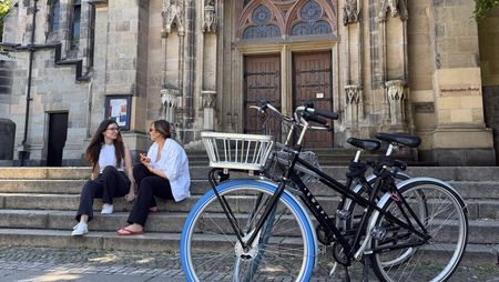 Zwei junge Frauen sitzen auf der Treppe vor Thomaskirche in Leipzig. Im Vordergrund stehen zwei Fahrräder von Swapfiets. 