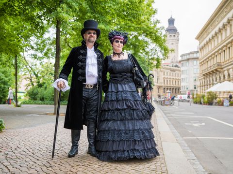 Links ein Mann in schwarzem Anzug und rechts eine Frau in schwarzem Kleid vor einem Park