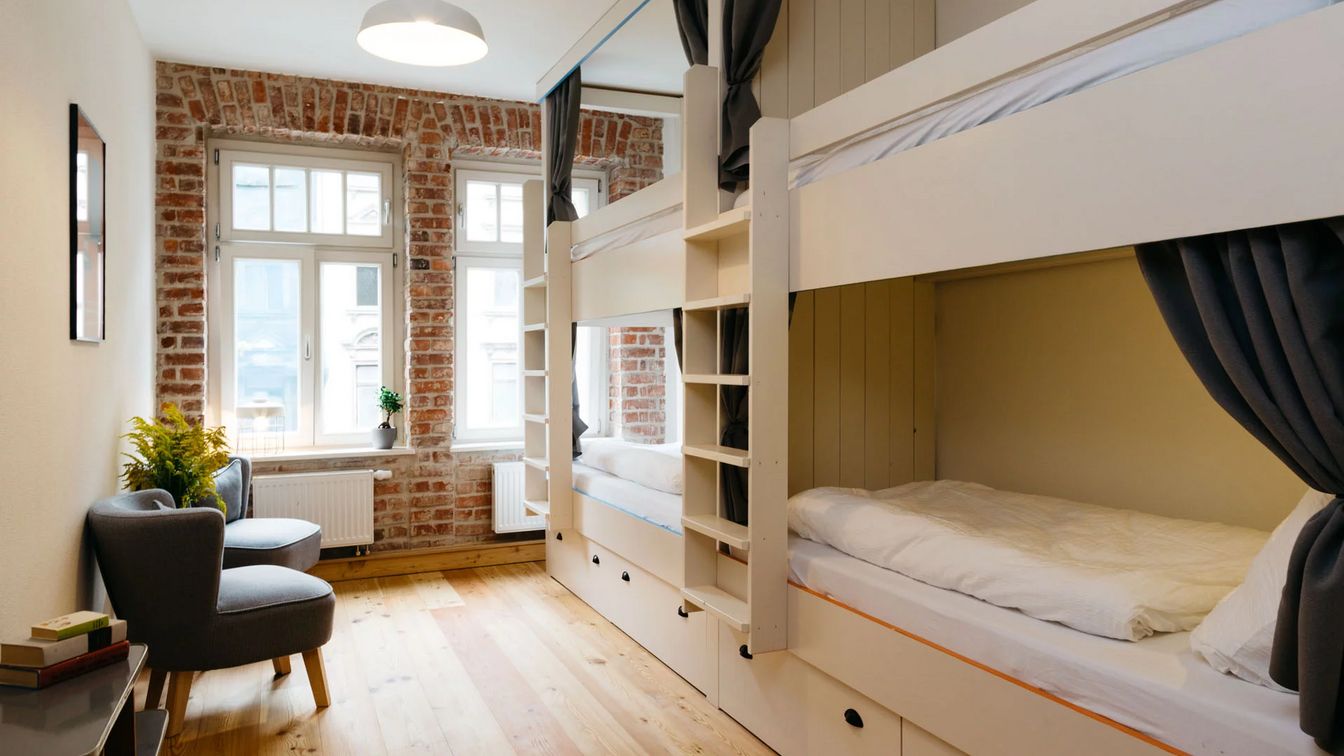 Blick auf ein helles und modernes Hostelzimmer mit zwei Etagenbetten und zwei Sesseln.