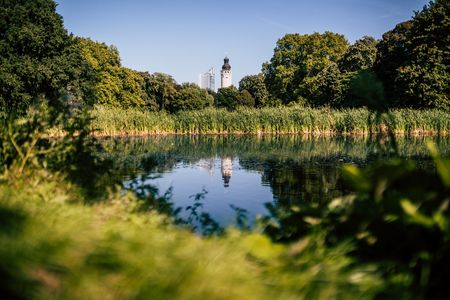 Die Türme des City-Hochhaus und des Neuen Rathaus ragen aus den Baumkronen hervor und spiegeln sich im Gewässer des Johannaparks