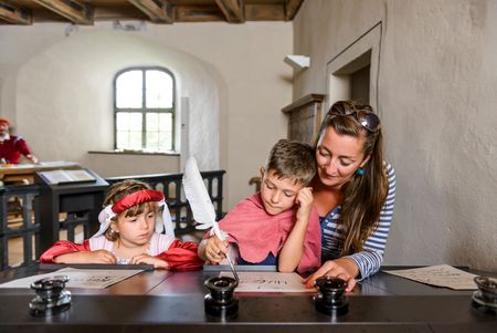 Eine Mutter sitzt mit ihren Kindern verkleidet als Prinzessin und Ritter an einem Tisch und hilft ihnen beim Schreiben mit Tinte und Feder.