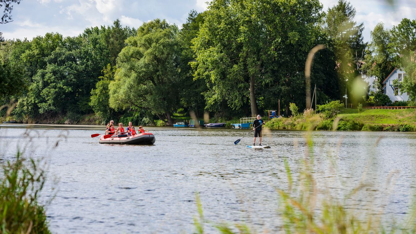 Eine Familie paddelt in einem Schlauchboot auf der Mulde in Richtung eines Mannes auf einem Stand-Up Paddle Board.